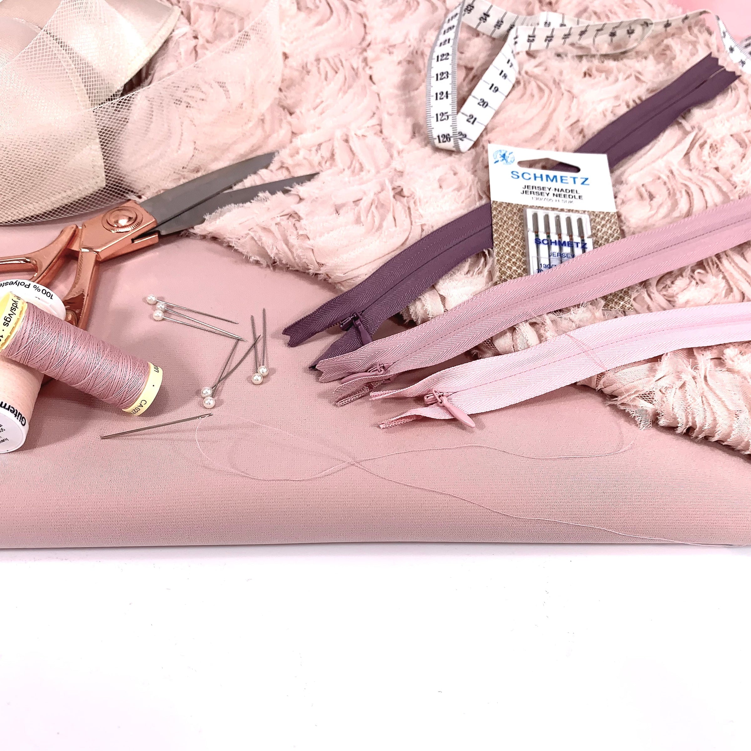 Flat lay of sewing supplies including spools of threads, scissors, invisible zippers, tape measure, sewing machine needles, and bra cups on pink textured fabric - haberdashery supplies from Fabric Collection Brisbane